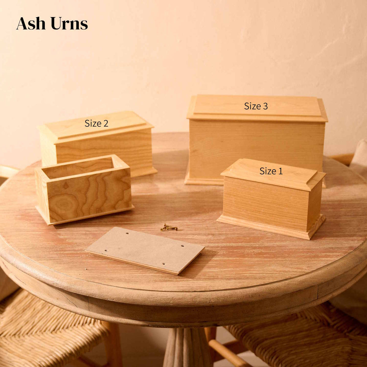 Four wooden cremation urns of different sizes on a round wooden table. The urns are all rectangular and are all made of Ash wood. they have bevelled edges. There are two identical small urns, which are labelled "Size 1". One of these small urns has its lid removed, showing the inside of the urn (which is empty). The lid and four screws sit on the table. The medium urn is labelled "Size 2". The large urn is lablled "Size 3". Both the medium and large urns are shown with their lids on.